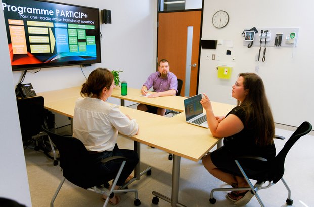 Trois personnes travaillant dans un laboratoire de simulation clinique avec équipements informatiques et médicaux. Les personnes sont assises autour d’une table et l’une d’entre elles a son portable et sa présentation est affichée sur le grand écran. 