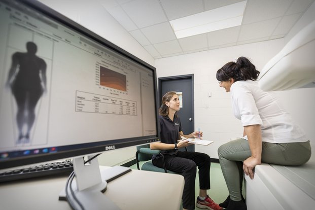 Dans une salle de consultation en kinésiologie, un écran avec un représentation du corps humain et, à l'arrière-plan, une étudiante qui discute avec une patiente dans.