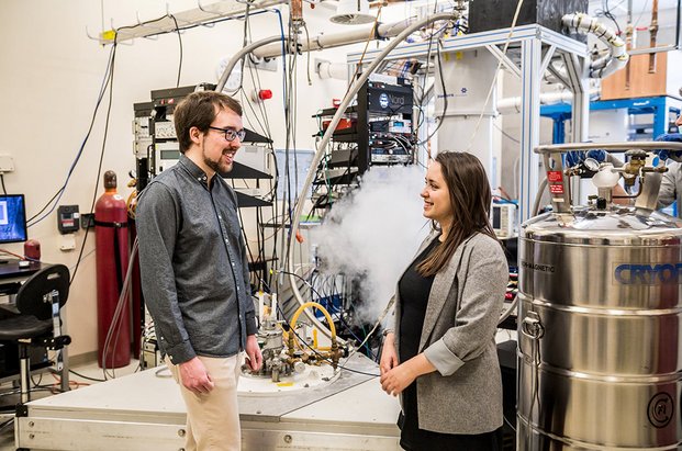 Un étudiant et une étudiante dans un laboratoire de physique quantique avec beaucoup d’appareils (cuve de refroidissement, écrans, filage.etc).