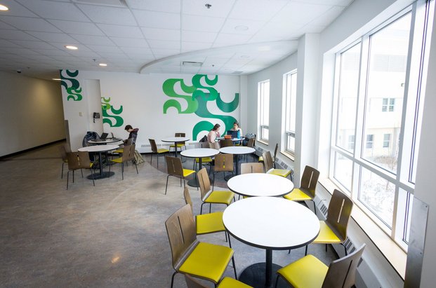 Petites tables bistro et chaises colorées dans un espace très fenestré avec mur du fond décoré du S de l'Université