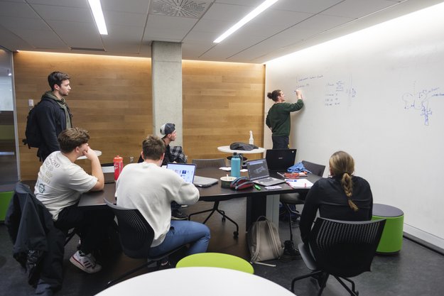 Personnes étudiantes à une table de travail en regardent une autre écrire sur le mur blanc.