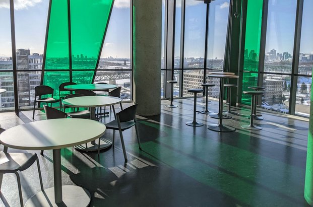 Grand espace avec une fenestration transparente et verte donnant une vue sur la ville de Longueuil et dans lequel sont disposées des tables et des chaises. 