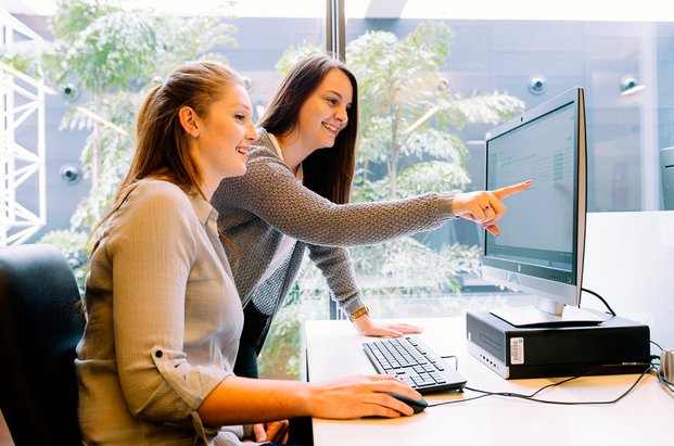 Deux personnes travaillant devant un ordinateur à la bibliothèque, l’une pointant l’écran avec son doigt pour montrer quelque chose à l’autre. 