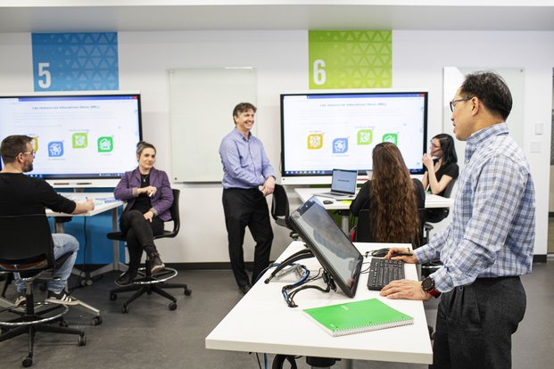 Des grands écrans informatiques sur le mur d'une salle avec des table de travail d'équipe et plusieurs personnes en discussion et réflexion