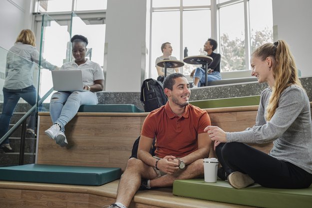 Personnes étudiantes en conversation, installées par groupes de deux de manière décontractée dans une salle avec bancs coussinés, en paliers, avec murs de fenêtres.