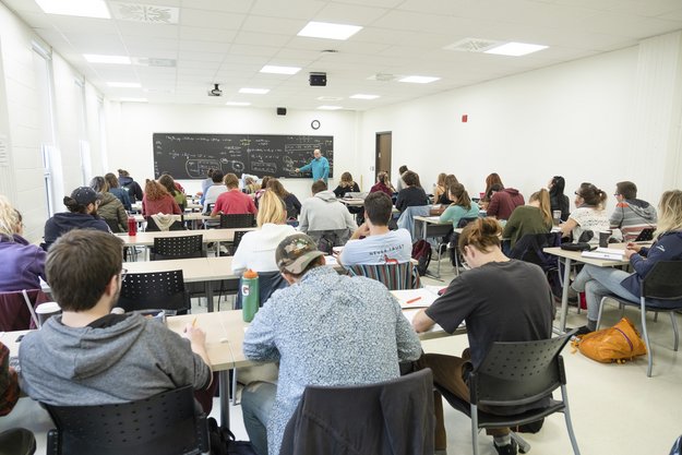 Classe remplie de personnes étudiantes à des tables qui écoutent le professeur, qu'on voit devant le tableau noir.