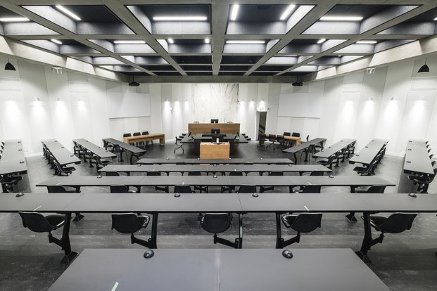 Salle aux murs blancs et mobilier noir structurée comme une salle d'audience judiciaire.