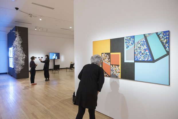 Une personne regarde de près un grand toile sur um mur. À l'arrière-plan, d'autres personnes circulent dans la galerie.