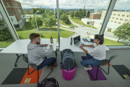 Un étudiant et une étudiante à un table de travail  devant un mr complètement vitré avec vue sur la verdure et les bâtiments du campus