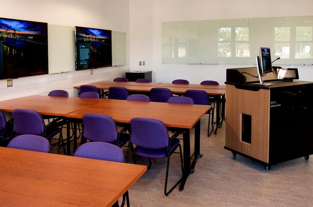 Salle de classe avec des tables, des chaises, des écrans et un podium. 