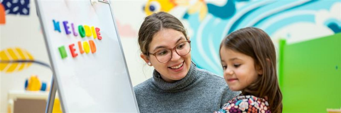 Enseignante aidant une petite fille d'âge préscolaire à placer des aimants alphabétiques de couleurs sur un tableau blanc